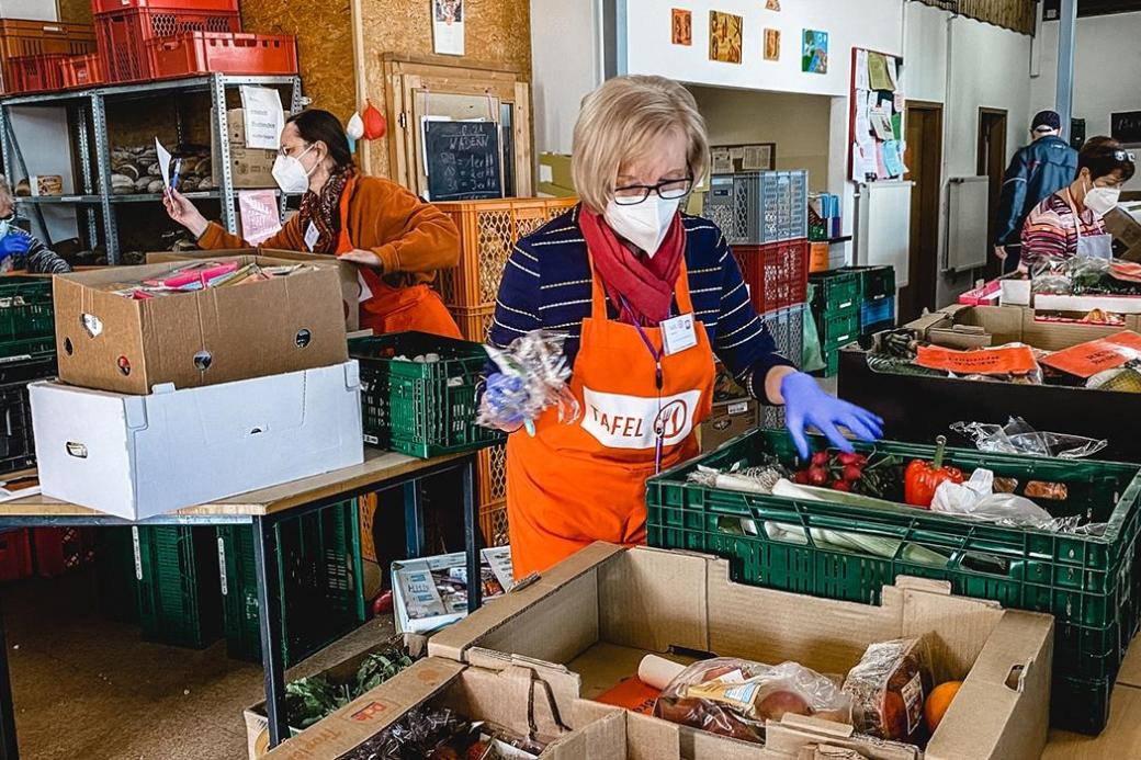 Inmitten Regalen von Lebensmitteln stehen zwei Menschen, die sortieren. Sie tragen Schützen mit der Aufschrift Tafel